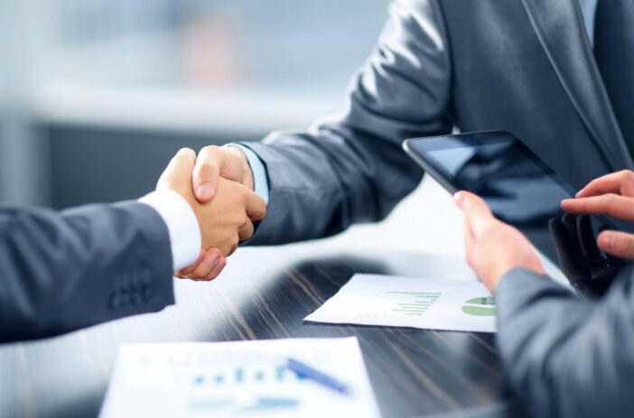 Two professional men shaking hands and a third man holding a tablet- 22928632